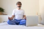 Mature adult man sitting on bed at home following online meditation using laptop and headphones.